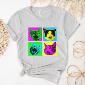 May include: A light gray t-shirt with a pop art design featuring four cats with different colored backgrounds. The cats are all looking at the viewer with their mouths open. The colors of the backgrounds are pink, blue, green, and yellow.