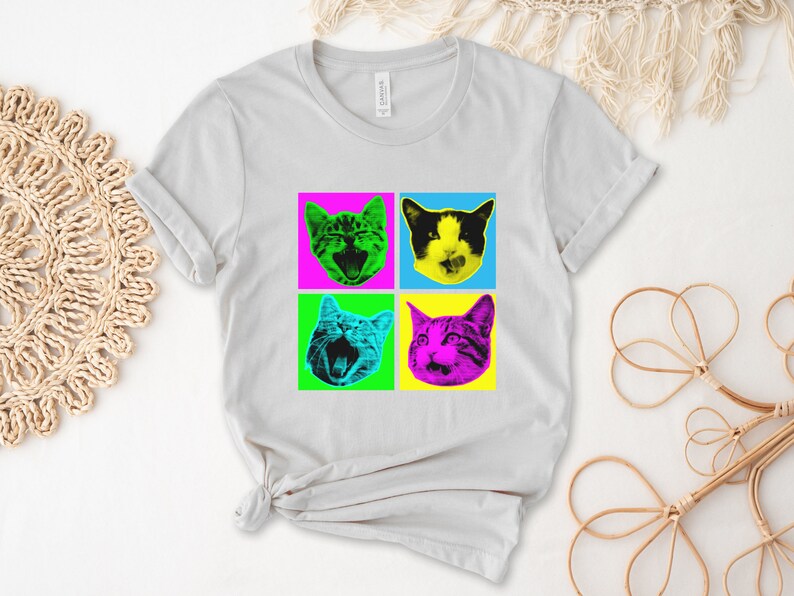 May include: A light gray t-shirt with a pop art design featuring four cats with different colored backgrounds. The cats are all looking at the viewer with their mouths open. The colors of the backgrounds are pink, blue, green, and yellow.