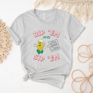 May include: A light gray t-shirt with the text "Rip 'Em and Sip 'Em" in pink and green. A cartoon beer mug with a smiling face is holding a lottery ticket.