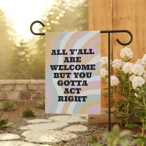May include: A colorful garden flag with a wavy pattern in shades of pink, blue, and yellow. The flag has the text "All y'all are welcome but you gotta act right" in black.