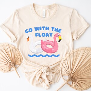 May include: A beige t-shirt with the text "Go with the float" printed on it. The text is in blue and there are two inflatable pool floats, a swan and a flamingo, in the image.
