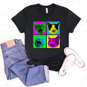 May include: Black t-shirt with a pop art design of four cats with different colored backgrounds. The cats are all looking at the camera with their mouths open.