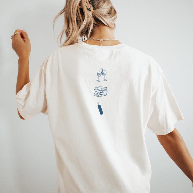 Op de afbeelding: Wit T-shirt met een blauwe afbeelding van twee wijnglazen die klinken, een stapel boeken en een bladwijzer.