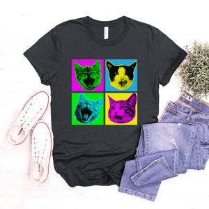 May include: A dark grey t-shirt with a pop art design featuring four different cat faces in colorful squares. The cat faces are in a grid pattern with a black background.