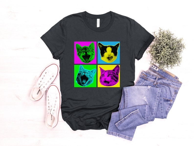 May include: A dark grey t-shirt with a pop art design featuring four different cat faces in colorful squares. The cat faces are in a grid pattern with a black background.