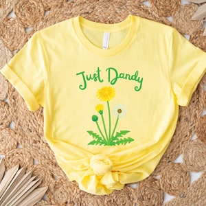 Op de afbeelding: Geel T-shirt met een groene afbeelding van paardenbloemen en de tekst "Just Dandy".