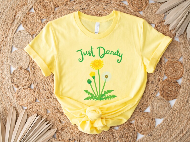 Op de afbeelding: Geel T-shirt met een groene afbeelding van paardenbloemen en de tekst "Just Dandy".