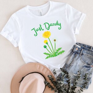 Op de afbeelding: Wit T-shirt met een groene afbeelding van een paardenbloem en de tekst "Just Dandy".
