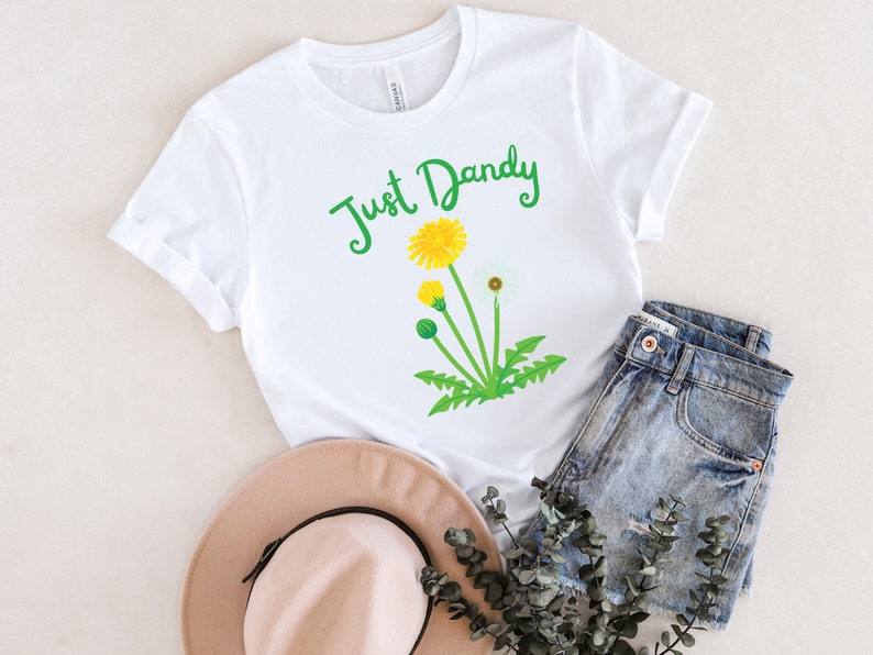 Op de afbeelding: Wit T-shirt met een groene afbeelding van een paardenbloem en de tekst "Just Dandy".