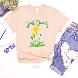 Op de afbeelding: Een beige T-shirt met een groene, gele en witte paardenbloem afbeelding en de tekst "Just Dandy" in groen.