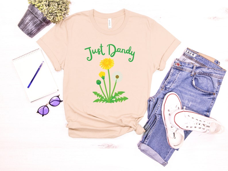 Op de afbeelding: Een beige T-shirt met een groene, gele en witte paardenbloem afbeelding en de tekst "Just Dandy" in groen.