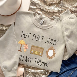 May include: A beige sweatshirt with the text "Put that junk in my trunk" printed on it. The text is in black and there are three illustrations of vintage items: a lamp, a radio, and a mirror.