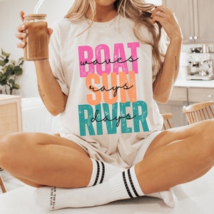 May include: A white t-shirt with a graphic design that says "BOAT waves SUN rays RIVER days" in colorful, distressed lettering.