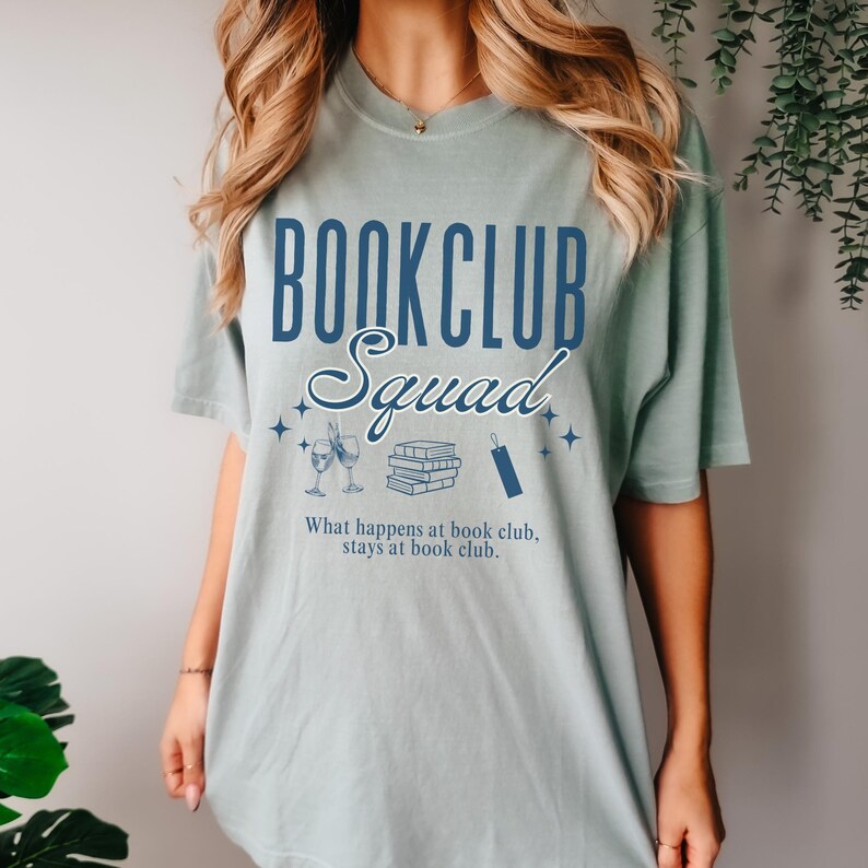 Op de afbeelding: Een lichtblauw T-shirt met de tekst "Bookclub Squad" in blauwe letters. Het shirt heeft ook een afbeelding van een stapel boeken, twee wijnglazen en een bladwijzer. De tekst "What happens at book club, stays at book club." staat onder de afbeelding.