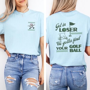 May include: Light blue t-shirt with a golf-themed design. The front features a cartoon golf ball character and the back displays the text "Get in LOSER We gotta find YOUR GOLF BALL" with a golf cart and flag.