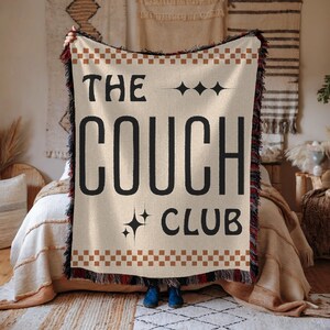 May include: A beige woven throw blanket with fringe edges features the text "The Couch Club" in black with a brown and white checkered border.