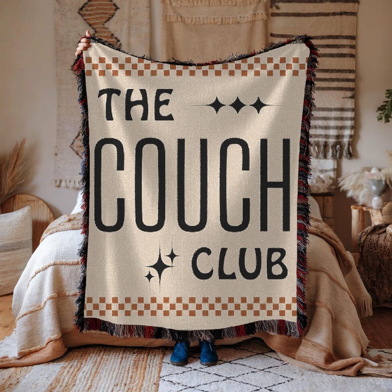 May include: A beige woven throw blanket with fringe edges features the text "The Couch Club" in black with a brown and white checkered border.