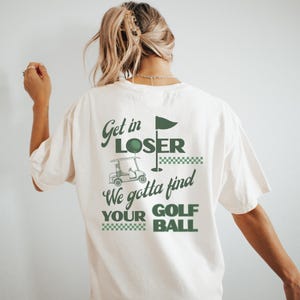 May include: Cream-colored t-shirt with a green golf-themed graphic. The design includes a golf cart, flag, and the text "Get in LOSER We gotta find YOUR GOLF BALL". The shirt is a relaxed fit.