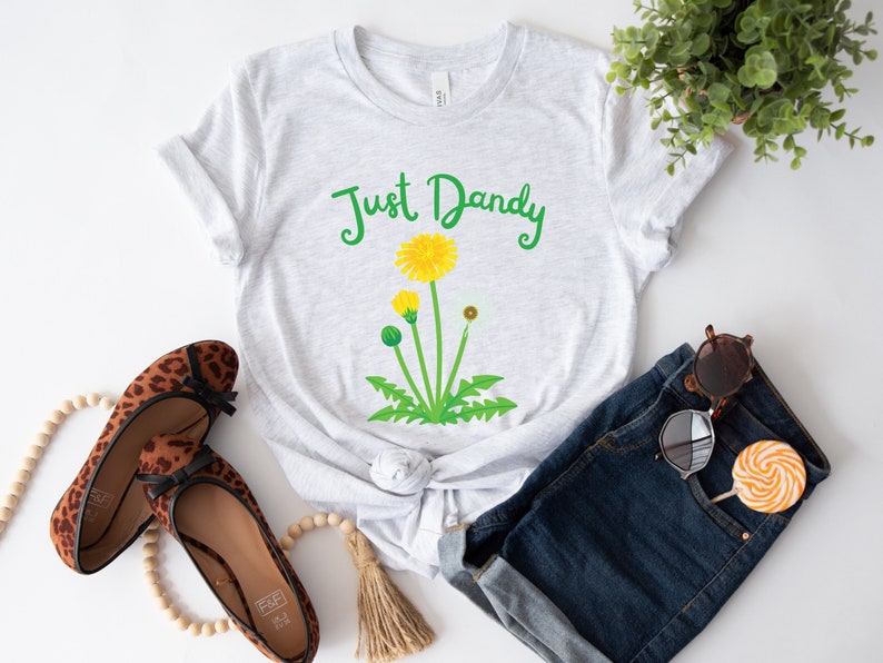 Op de afbeelding: Een grijs T-shirt met de tekst "Just Dandy" in groene letters en een gele paardenbloem afbeelding.