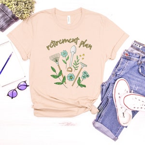 May include: A beige t-shirt with the text "retirement plan" in black lettering. The shirt features a graphic of gardening tools, including a shovel, rake, and trowel, with green leaves and blue flowers.