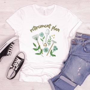 May include: White t-shirt with the text "retirement plan" in gold lettering. The design features a rake, shovel, trowel, and gardening gloves with green leaves and blue flowers.