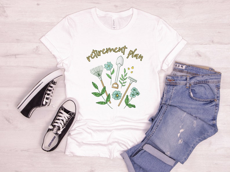 May include: White t-shirt with the text "retirement plan" in gold lettering. The design features a rake, shovel, trowel, and gardening gloves with green leaves and blue flowers.