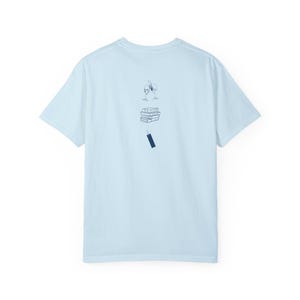 Op de afbeelding: Lichtblauw T-shirt met een afbeelding van een stapel boeken, een glas wijn en een bladwijzer.
