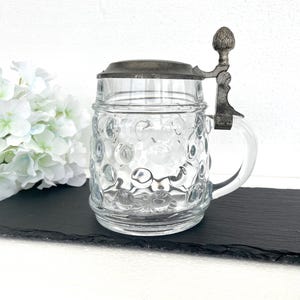 May include: Clear glass beer stein with a metal hinged lid and handle. The stein has a textured, bubble-like pattern on the glass. The metal lid and handle have an aged, antique appearance. The stein is displayed on a black surface.