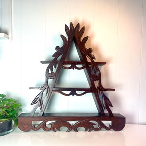 Puede incluir: Estantería de madera marrón oscuro con diseño en forma de A. La estantería tiene tres niveles y está adornada con patrones florales y de hojas talladas. La base y la parte superior del marco también presentan tallas decorativas.