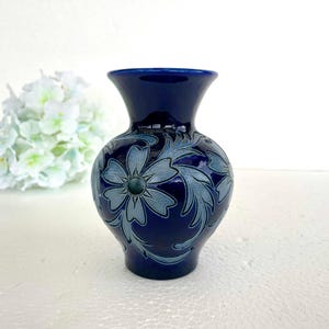 Peut inclure: Un vase en céramique bleu foncé avec une finition brillante. Le vase présente un motif floral dans des tons bleu clair. Le vase a un corps arrondi et un bord évasé. C'est un objet décoratif.