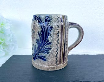 May include: A stoneware beer stein with a handle. The mug features a blue floral design on a beige background. Vertical brown and beige stripes are on the side. The handle is a darker shade of brown. The stein is sitting on a black surface.
