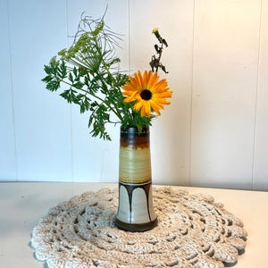 May include: A ceramic vase with brown, white, and yellow glaze is filled with a single orange flower and green foliage.