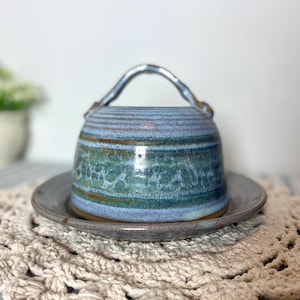 Op de afbeelding: Een blauwe en bruine keramische botervloot met een handvat. De vloot heeft een geglazuurde afwerking en staat op een gehaakt onderzetter.