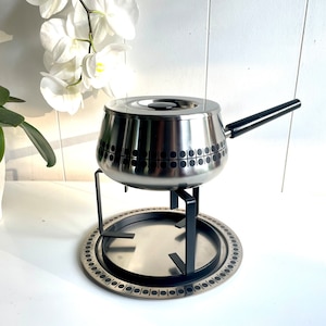 May include: A silver fondue pot with a black handle and a geometric pattern sits on a black metal stand with a silver tray. The stand has a decorative pattern of circles around the edge.