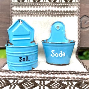 Può includere: Due contenitori vintage in metallo blu chiaro, uno con la scritta "Salt" e l'altro "Soda". Entrambi hanno un aspetto invecchiato e rustico con un anello per appenderli in alto. I contenitori sono appoggiati su un tessuto fantasia.
