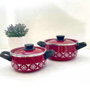 May include: Two red enamel pots with white floral designs and black handles. The smaller pot is in the foreground and the larger pot is in the background.