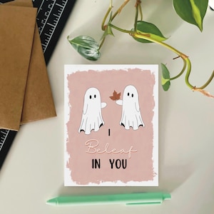May include: A rectangular greeting card with a dusty pink background featuring two white cartoon ghosts. One ghost holds a brown autumn leaf. The text reads "I Beleaf IN YOU" in a mix of cursive and block lettering. The card has a white border.