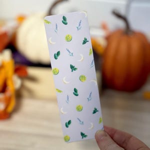 May include: A rectangular bookmark with a light purple background. The bookmark is decorated with watercolor illustrations of green lime slices, mint leaves, lavender sprigs, and crescent moons. The bookmark is held by a person.