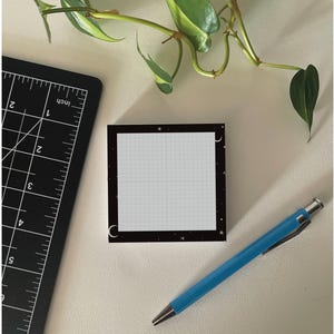 May include: A square sticky note with a light gray grid pattern on a white background, framed by a black border featuring white stars and crescent moons. A black ruler with white inch markings and a blue mechanical pencil are positioned nearby.