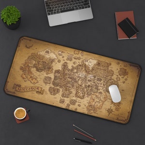 May include: A large rectangular desk mat with a detailed map design, featuring the word "Gielinor" in a banner. The mat is brown with a black border, and a white mouse rests on the surface. A laptop, notebook, and pencils are also visible.