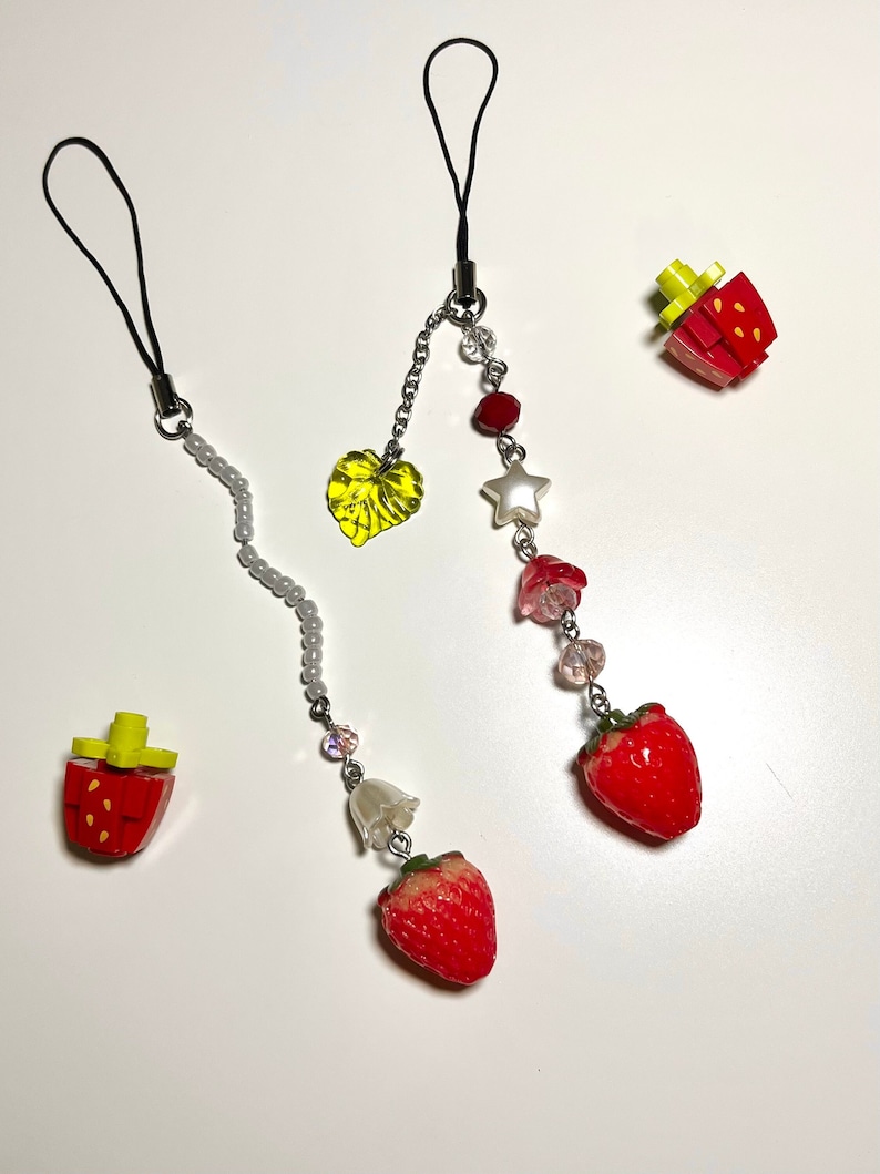 May include: Two phone charms with a strawberry, leaf, star, and flower design. One charm has a white pearl chain and the other has a silver chain.