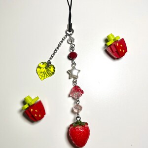May include: A red and green strawberry charm with a silver star, beads, and a green leaf charm hanging from a black phone strap.