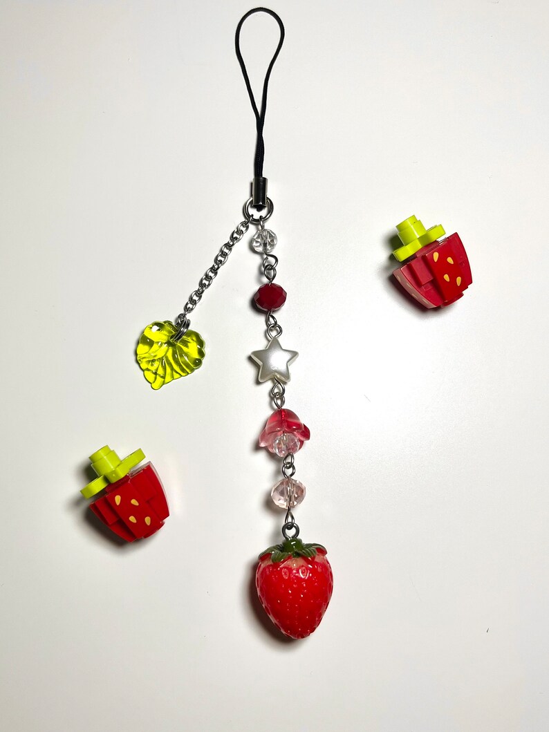 May include: A red and green strawberry charm with a silver star, beads, and a green leaf charm hanging from a black phone strap.