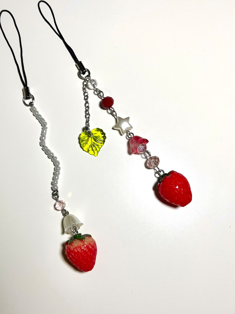 May include: Two phone charms with a strawberry, leaf, and beads. One charm has a white bell and the other has a star and pink flower.