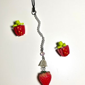 May include: A gray beaded phone charm with a white bell and a red strawberry charm. The charm is attached to a black string.