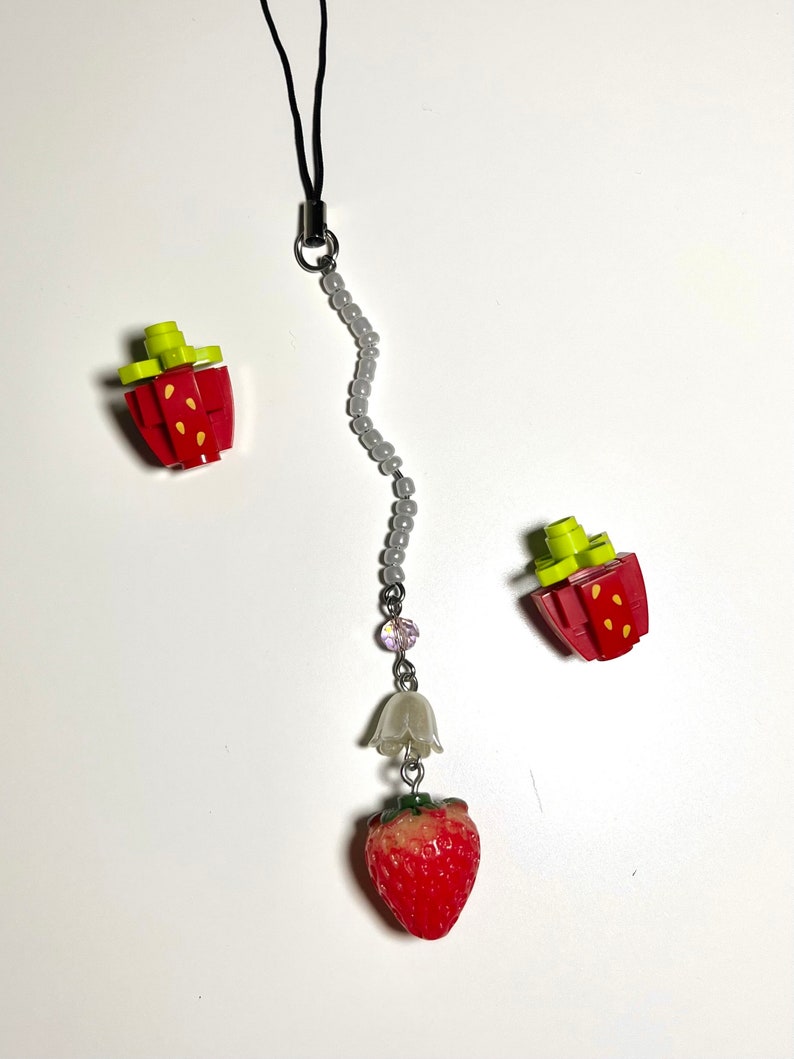 May include: A gray beaded phone charm with a white bell and a red strawberry charm. The charm is attached to a black string.