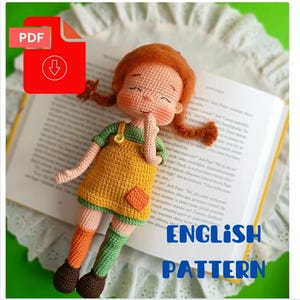Puede incluir: Una muñeca de ganchillo hecha a mano con cabello rojo, que lleva un vestido amarillo con mangas verdes y calcetines naranjas y verdes. La muñeca está acostada sobre un libro abierto. La imagen incluye un icono PDF rojo con las palabras "English Pattern".