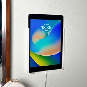 May include: A black tablet is mounted on a white wall-mounted holder. The tablet displays a colorful wallpaper with the time 18:35 and the battery status. A white charging cable is plugged into the tablet.