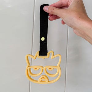May include: A yellow cat-shaped pancake mold with a grumpy face design, featuring a black strap with a yellow button. The mold is held up against a white background.