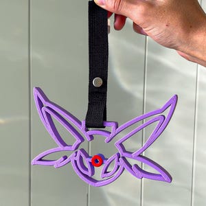 May include: A purple, stylized, butterfly-shaped object with a black strap and a silver button. The butterfly has a red circle in the center. The object is held by a hand.